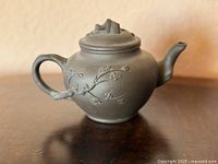 Left side view of dark clay teapot showing handle, spout and floral relief