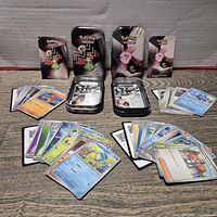 Overview of common Pokémon cards spread around two open metal tins with additional tin lids/packaging behind