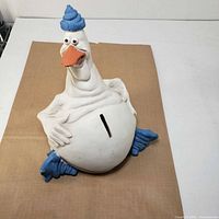 Front view of white resin goose piggy bank with blue hat and feet, coin slot on belly visible