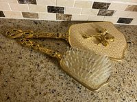 Hand mirror and brush angled, showing gold-tone frames and bristles