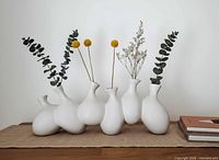 Front view showing six connected white ceramic bud vases with stems for scale