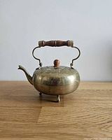 Side view of brass teapot on feet with wooden swing handle and spout visible