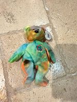Tie-dye Peace Beanie Baby bear with tag protector on stone floor