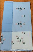 Folded tablecloth showing blue and white panels with pink floral embroidery