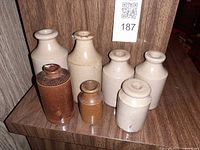 Front view of seven stoneware ink bottles (five off-white, two brown) on shelf