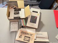 Top-down view showing Commodore C2N unit in box with foam, cable, multiple cassette tapes, Zygon booklets and instruction sheets