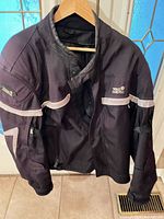 Front view of black Tour Master armored motorcycle jacket hanging, showing reflective piping and logo