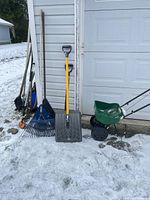 Group of assorted garden tools and spreader leaning against garage