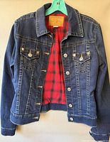 Front view of dark blue denim True Religion jacket on hanger showing red plaid lining, chest pockets, button front