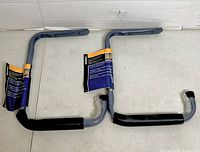 Two grey metal shelf brackets with labels attached, black foam on bottom arms