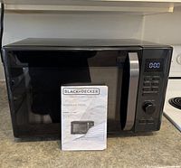Front view of Black+Decker microwave oven showing door, handle, control panel and included instruction manual
