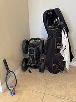Black RAMPION golf bag with clubs, folded Caddytek push cart, AMF blue racquetball racquet with ball and umbrella visible