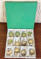 Open green display box with 12 rotary dial pendant necklaces in individual sleeves