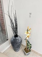 Large grey vase with black branches and adjacent orchid in blue planter