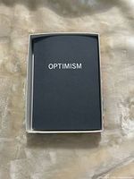 Black hardcover journal inside box showing title "OPTIMISM"