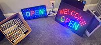 LED "OPEN" and "WELCOME OPEN" signs lit, two monitors, box of DVDs and CDs