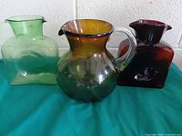 All three glass pitchers: green square, amber-to-green round with handle, ruby square