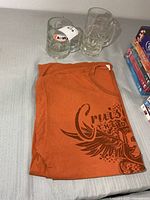 Folded orange A&W graphic T-shirt beside two clear glass logo mugs