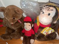 Overview showing all three plush toys including bagged 25 in Curious George, small Curious George and Gund teddy bear