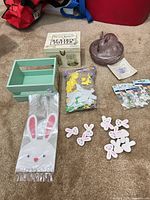 All items arranged on carpet: boxed book set, purple glass bunny dish (lid off), green crate, sealed foam shapes, wooden bunny cut-outs, treat bags pack, two sticker packs