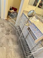 Overview of three white wire racks, wooden 3-drawer organizer and tabletop vanity mirror in bathroom setting