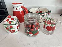 Group shot showing pitcher, sugar jar, glass creamer, decorated glass jar with wooden cherries, ceramic colander, matching plate