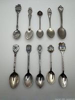 Ten assorted souvenir teaspoons laid out in two rows showing decorative handles and enamel crests