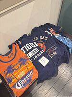 All four shirts laid out—orange Corona tank plus three navy tees (Antigua, Air Jordan, Black Keys)