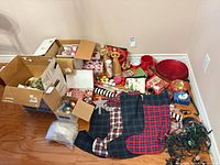 Overview of entire mixed lot on floor showing boxes of ornaments, stockings, wooden figures, red bowls and more