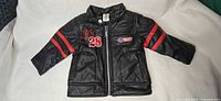 Front view of black baby faux leather jacket showing zipper, red sleeve stripes, M28 embroidery and Moto patch