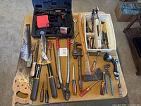 Overall view of tools on table including drill, hand saw, lopper, pipe wrench, axe, screwdrivers, chisels, wrenches and tote
