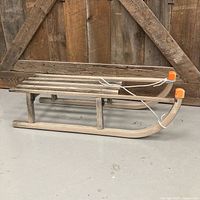 Side view of wooden sled displaying slatted seat, curved runners, orange tip caps