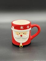 Front view of red ceramic mug with Santa face relief, snowflakes on rim, black belt at base