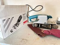 Makita 9924DB belt sander with dust bag, sanding belt and box on shelf