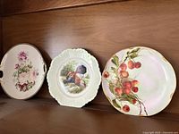 Three plates on shelf: pink roses plate, turkey scene plate, cherry design plate