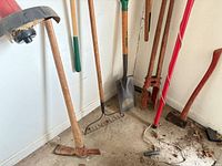 Group of long-handled garden tools standing against wall: mattock, bow rake, shovel, post-hole digger, splitting maul, manual string trimmer