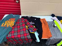 Six clothing items laid out including teal tee, plaid dress, Lakers tee, orange shorts, grey shorts, neon yellow pants