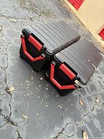 Two black hard-sided plastic totes with red latches on paved surface