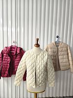 Front view of three puffer jackets in magenta, cream and beige on hangers and mannequin