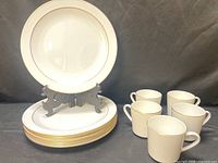 Stack of eight dinner plates with gold rim and five coffee cups displayed