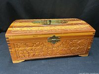 Three-quarter view of carved wooden jewelry box showing front floral panel, brass-tone latch, curved lid, block feet