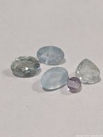 Five loose gemstones on white background—four light blue aquamarines and one purple bead