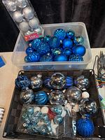 Clear tub filled with assorted blue ornaments and package of silver ornaments; woven basket of mixed blue and silver bulbs