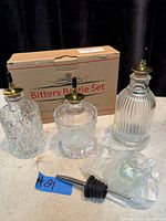 overview of three glass bitters bottles with installed spouts, extra spout in bag, original box visible