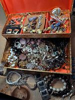 overview of open two-tier box showing upper tray with earrings, brooches, lower compartment filled with assorted jewellery and items arranged outside the box