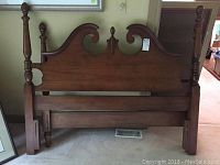 Solid wood double size headboard with ornamental carved scrolling and turned finials on posts, shown standing against a wall.
