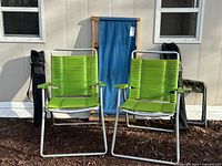 Front view of two green vinyl aluminum chairs, blue fabric wooden lounger behind, camping chairs in black bags at sides, camo stool partially visible