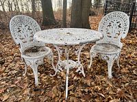 Full view of ornate white painted cast aluminum patio table and two matching chairs on leaf-covered ground