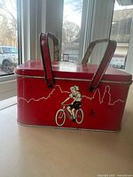 Side view of red tin lunch box showing girl on bicycle graphic and two metal handles