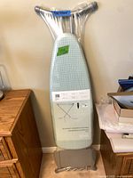Full ironing board leaning against wall, packaging intact